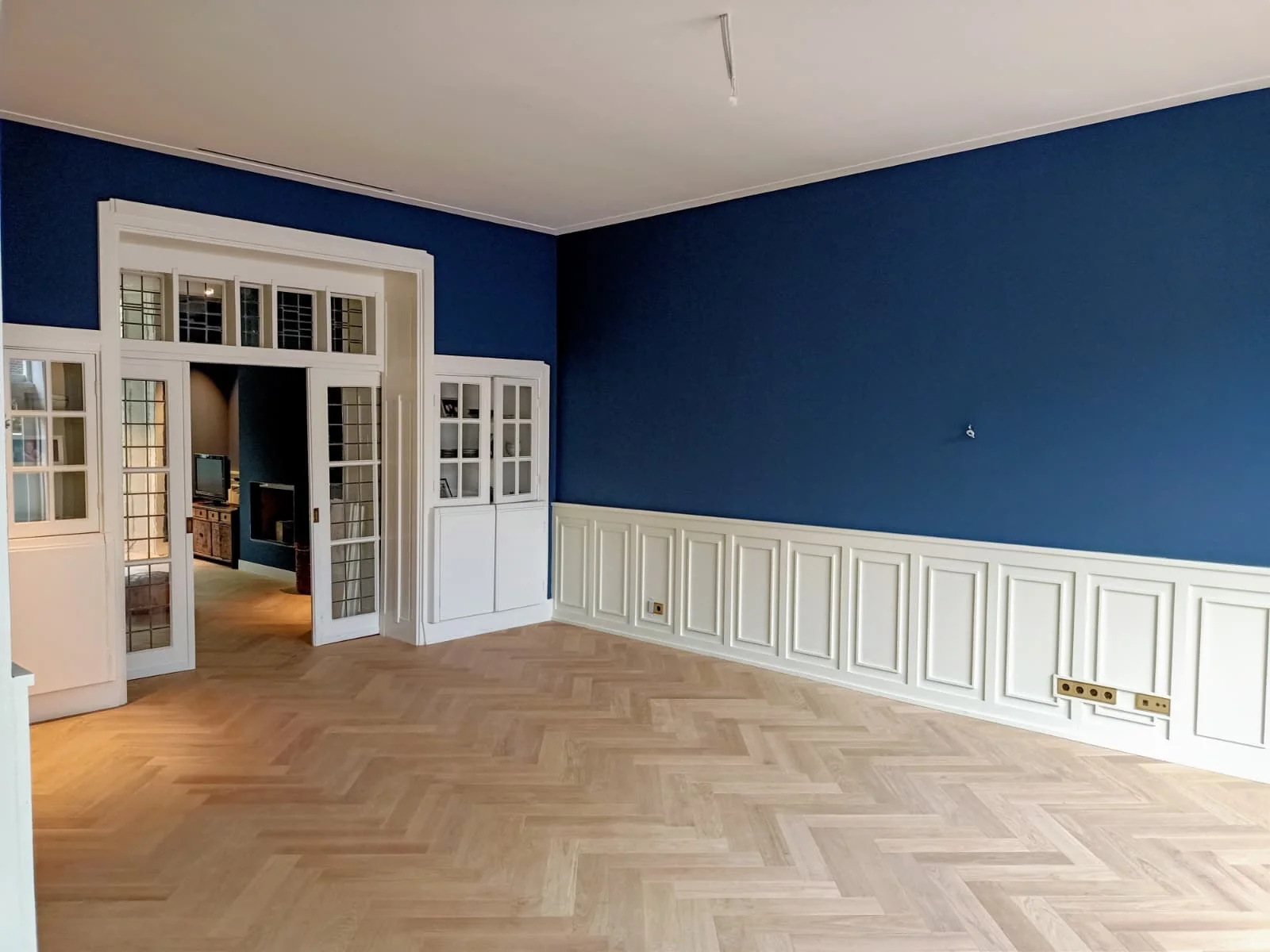 Een ruime, lege kamer met diep donkerblauwe muren boven wit lambrisering. De vloer is lichtgekleurd hout gelegd in visgraatpatroon. Links openen grote witte openslaande deuren met glaspanelen naar een aangrenzende kamer waar een open haard zichtbaar is. Witte ingebouwde kasten met glazen deuren staan naast de deuropening.