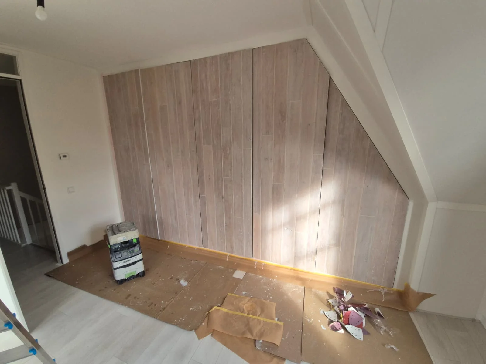 Een interieurfoto van een kamer die wordt verbouerd, met een grote accentmuur bedekt met verticale lichtgekleurde houtlook panelen die de schuine lijn van het plafond volgen. De vloer is beschermd met bruin karton, waarop een stapel schildersapparatuur en een hoop afval van behangpapier ligt. Links leidt een deuropening naar een hal met een wit traphek.